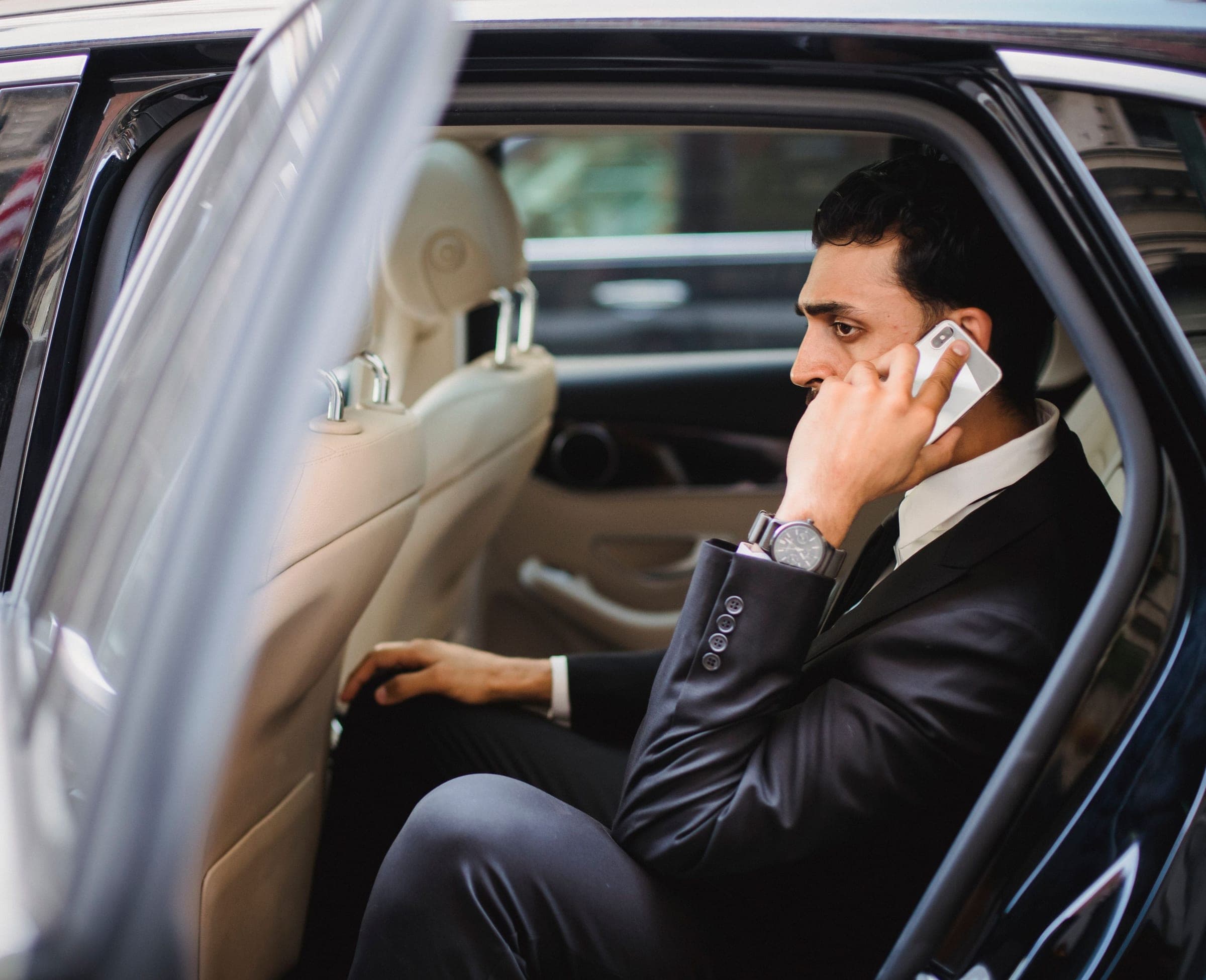 Executive taking a call in the rear seat of a black chauffeured sedan with the door held open