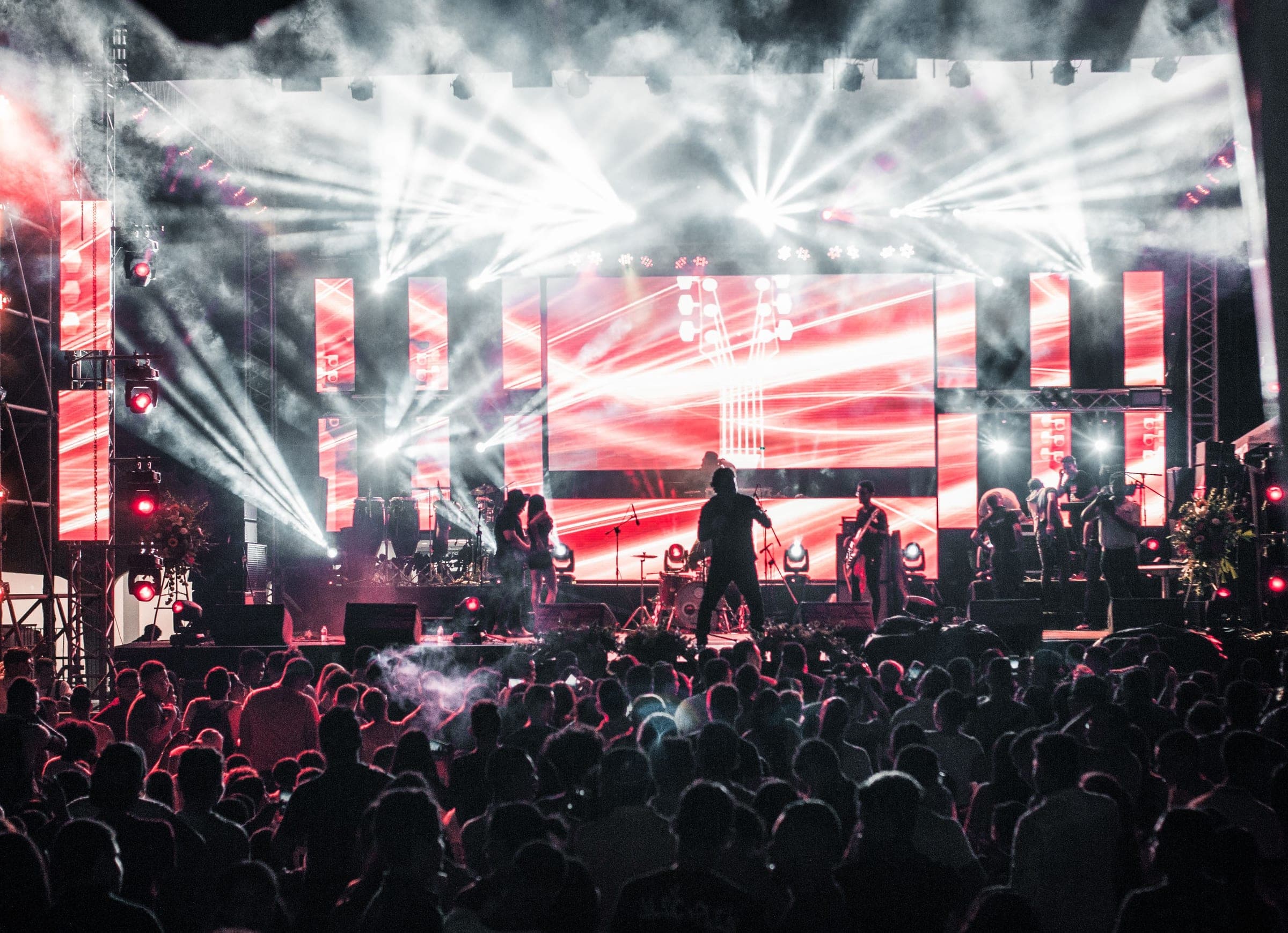 A festival stage lit in red and white at night, the crowd in silhouette in front of it