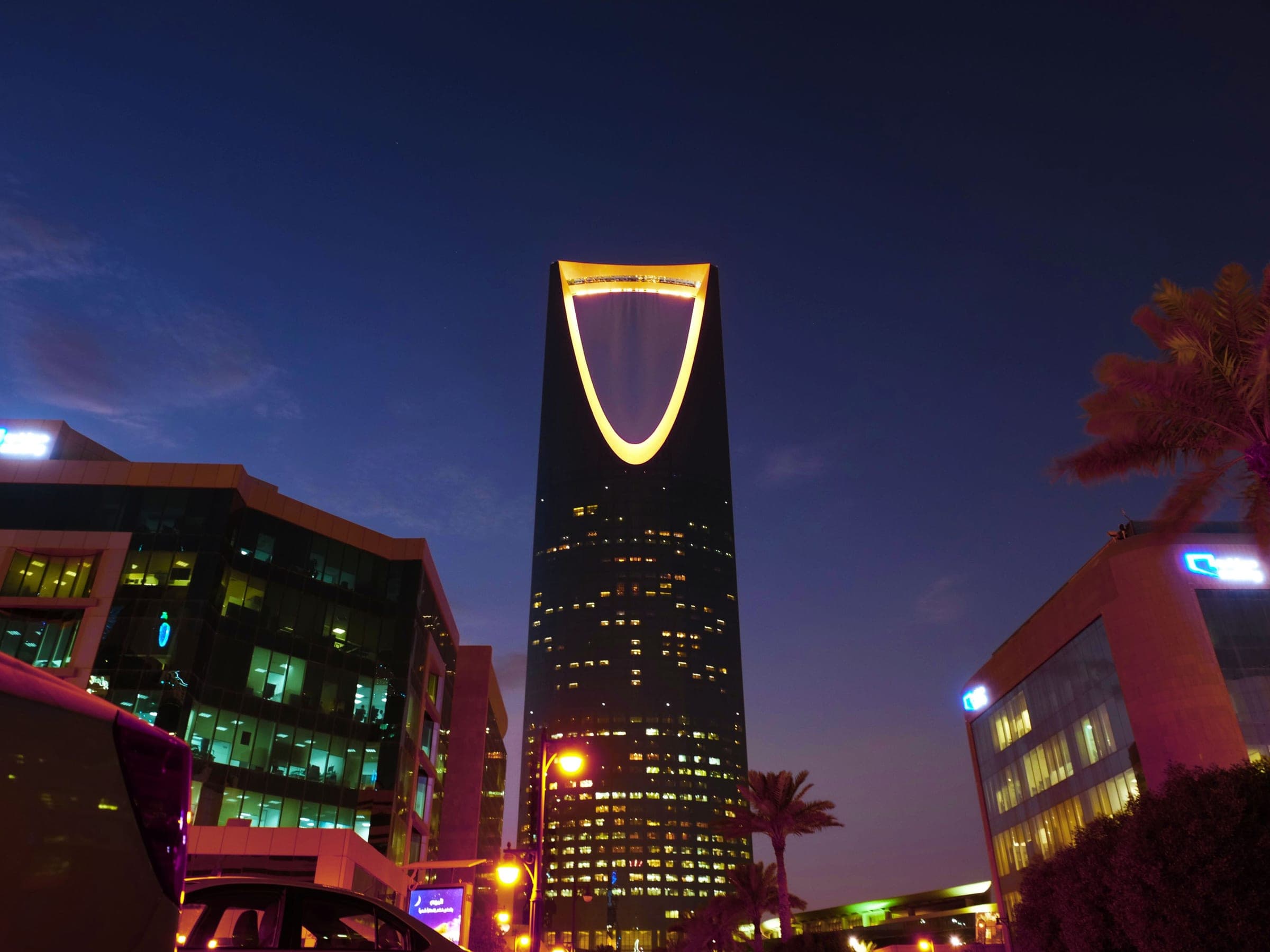 The Kingdom Centre lit at dusk above the office blocks and palms of Olaya in Riyadh, the district a held car spends a working day in